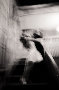 Black and white film photo of a newlywed couple sharing a kiss on the stairs of their wedding venue -  a candid moment captured by a film wedding photographer on black and white 35mm film. 