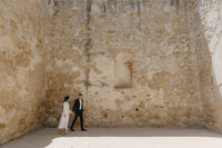 Engaged couple walking hand-in-hand along a dramatic, textured stone wall at Canopy Grove in Escondido, California.