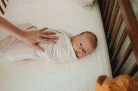 A baby wrapped in a white blanket is placed on a white mattress with wooden bars. A hand gently touches the baby.