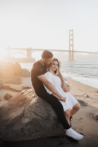 Black dressed man leaning against rock and woman in white dress leans in. Golden Gate Bridge in back at sunset