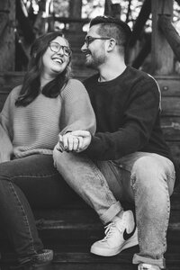 Lexi & Jared sitting on wood stairs laughing at each other as they hold out their hands to show off the engagement ring