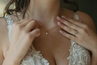 Bride adjusting delicate gold necklace featuring a single diamond pendant, captured in a close-up detail shot at Stone Spring Farms, Pennsylvania by a Destination Elopement Photographer — showcasing natural nails, elegant ring, and lace wedding gown.