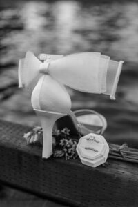 White bridal shoe with delicate bow detail displayed next to wedding rings in a hexagonal box, surrounded by soft floral accents on a rustic wooden surface, captured in a styled wedding detail shoot by a Portland and destination wedding photographer