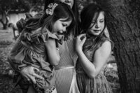 a black and white image of a mother holding her younger daughter and her older daughter is at her side in a gentle embrace.  The girls are looking down.  The mother's face cannot be seen.