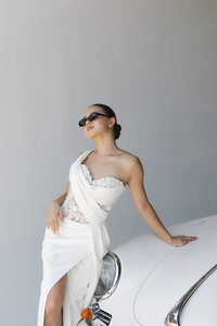 A stylish woman in a flowing white dress leans against a pristine white car, showcasing a chic and modern aesthetic.