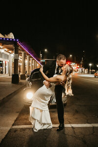 A joyful bride and groom celebrate in the middle of a crosswalk in Downtown Chandler. The groom throws up a hand in excitement while the bride laughs, holding onto his arm. Palm trees, traffic lights, and local buildings frame the lively city backdrop.