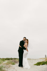 Bride in a strapless gown and groom in a black suit hugging on a sandy beach trail with coastal plants and grey skies.