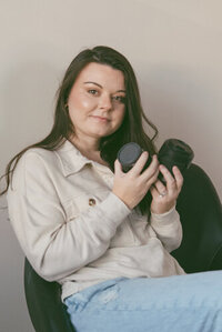 Client of Bostron Design Studio, Michela sits back in a chair in her office space, holding her camera and softly smiling.