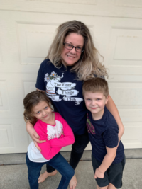 Woman poses with two smiling children in front of a neutral backdrop on cement.