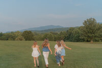 Four senior girls run playfully through a field with trees and mountains in the background in Northeast, TN at Willow Springs Park.