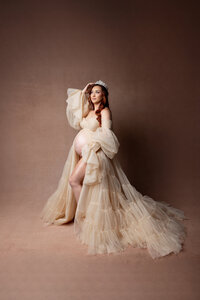 Expecting mom in a flowing champagne-colored tulle gown standing barefoot against a warm-toned studio backdrop. Photograph by Yucaipa maternity photographer Kaitlyn Dawn Photography.