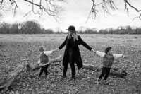 A mother wearing a long black wool coat and black hat stands with her young twin sons. Each holding her hands and pulling in the opposite direction. The photo is black and white against the winter backdrop of Bushy Park.