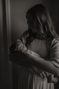 A black and white photo of a new mother holding her first child. Her face is in deep shadow while the light from the window reflects off the cheeks of a sleeping newborn babe.