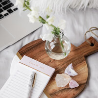 A notepad and pen, small glass vase with white flowers, and three crystal chunks sitting on a beautiful round wood charcuterie board with an open laptop to the side.
