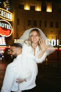 A couple celebrates their marriage by posing on the vibrant streets of Las Vegas at night, with the neon lights of casinos glowing in the background. One partner, wearing a veil, is playfully lifted by the other.