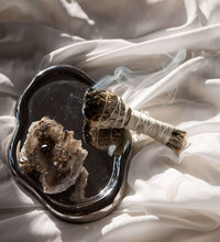 Sage and a crystal on a reflective tray with smoke rising, representing spiritual cleansing.