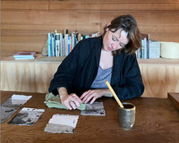 Solange Roberdeau focuses on crafting Japanese paper, gently pressing sheets on a wooden table, with a traditional Japanese brush and mortar beside her, and a bookshelf in the background, conveying a serene creative atmosphere.