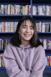 A young Asian teenager with dark, shoulder-length hair wears a purple hoodie and smiles over their left shoulder, past the viewer. The background of the image is a dark blue bookshelf, each shelf lined full of books, though the titles aren't visible.