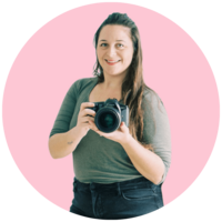 Sara, wearing a green shirt and black pants, holds her camera in her hands and smiles.