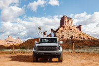 A couple is standing in their Bronco with the roof down waving their arms in excitement on their elopement day. They are standing in front of the Ida Gulch Trail.