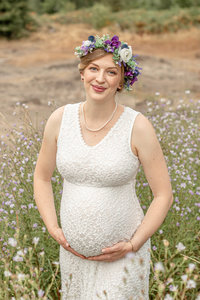 Woman in nature wearing a white lace maternity dress for a baby bump session. with a floral crown. Earth Mama Floral Pregnancy Photo Shoot