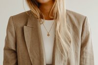 A close-up of a tanned blonde woman wearing a beige blazer over a white top. A gold necklace rests against her, with some of her hair tucked inside the blazer while the rest falls naturally.