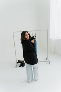 Photographer looks over her shoulder while holding a camera in front of a clothing rack in a naturally lit studio.