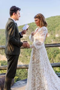 A bride and groom standing holding hands at the top of a mountain overlook. The groom is reading his vows to the bride while she has a hand on her chest and is holding back tears. 