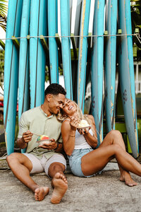 Playful couple sitting against stacked surfboards in Kauai, laughing and sharing Hawaiian shave ice during their engagement session.
