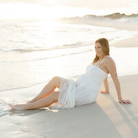 Maternity photo of a glowing mother sitting at the shoreline in a white dress, bathed in golden hour light along the coast.