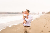 On Baker Beach in San Francisco dad is standing holding his toddler daughter and kissing her cheek while she smiles