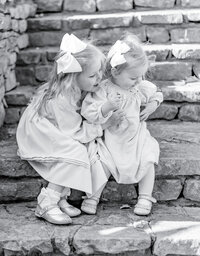 Black and white portrait of two young sisters sitting together on stone steps, dressed in classic smocked dresses with matching white bows. The older sister leans in with a smile, capturing the timeless beauty of sibling bonds in a heartfelt Birmingham family photography session