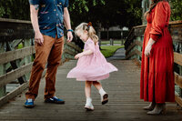 First Image (Family on Bridge): A young girl in a pink dress is twirling on a wooden bridge, her dress flowing around her as she spins. Her father, in a blue shirt and brown pants, and her mother, in a long red dress, watch her with smiles. The scene is set outdoors, with the bridge leading to a green, tree-filled park.