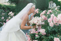 bride wearing a tiara and veil stops to smell a bush of pink roses in the garden at luxury cotswold wedding venue euridge manor