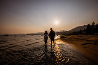 Bride and groom walking hand in hand along the shoreline of North Lake Tahoe at sunset, with golden light reflecting on the water.