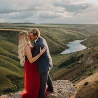 A man dressed in a nice suit holds the face of his girlfriend or fiance and kisses her. She is wearing a bright red dress and is holding his arm. Behind them is a valley with a river winding through it.