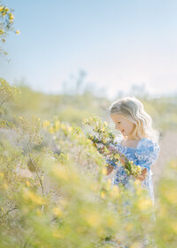 Photo of a blonde young girl wearing a blue and white dress  delicately holding yellow flowers amidst a field of flowers on a sunny day.