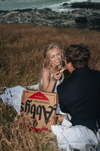 Newlywed couple sitting in the grass along the Oregon Coast during their summer elopement, as the bride smiles while feeding the groom pizza from delivery boxes.