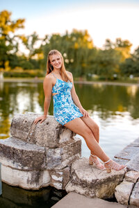A gorgeous blonde haired girl in a tight blue and white dress leaning against a stone structure with a pond behind her in Tower Grove park in St. Louis, MO.