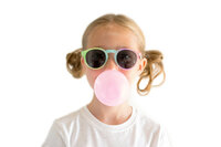 Girl blowing a pink bubble wearing rainbow sunglasses on a white backdrop