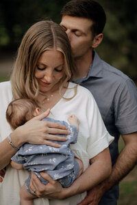 Dad stands behind Mom and gives her a kiss while Mom holds their newborn baby close.