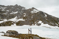 A couple stands together beside a frozen alpine lake surrounded by rugged, snow-covered mountains during their Alaska elopement, captured by Sydney Breann Photography.