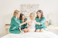 A bride in a white robe sits on a bed toasting champagne with her bridesmaids in green pajamas