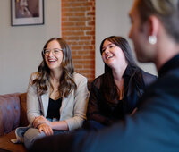 Two women laugh while seated on a leather couch during a casual meeting at SoHo63, a Chandler wedding and event venue. Exposed brick and framed art decorate the space as they chat with another guest, creating a warm and welcoming atmosphere.