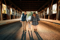 Parents walking with toddler under a covered bridge in front of sunset at golden hour