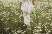 Woman in white dress walks through grassy field filles with white flowers.