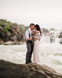 A newlywed couple poses for a stunning portrait at the water's edge during a session at Great Falls National Park in Virginia, captured by Nathan Sottung Photography after hiking to the base of the falls.
