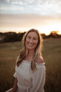 Blonde woman  looking right at the camera, she is wearing a long white skirt with a an off the shoulder matching top. She is on the beach at sunset.