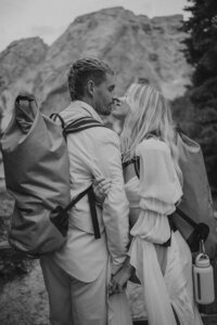 A bride and groom kiss in the mountains with hiking backpacks on