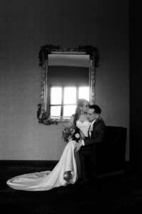Bride sitting on groom's knee in front of a mirror